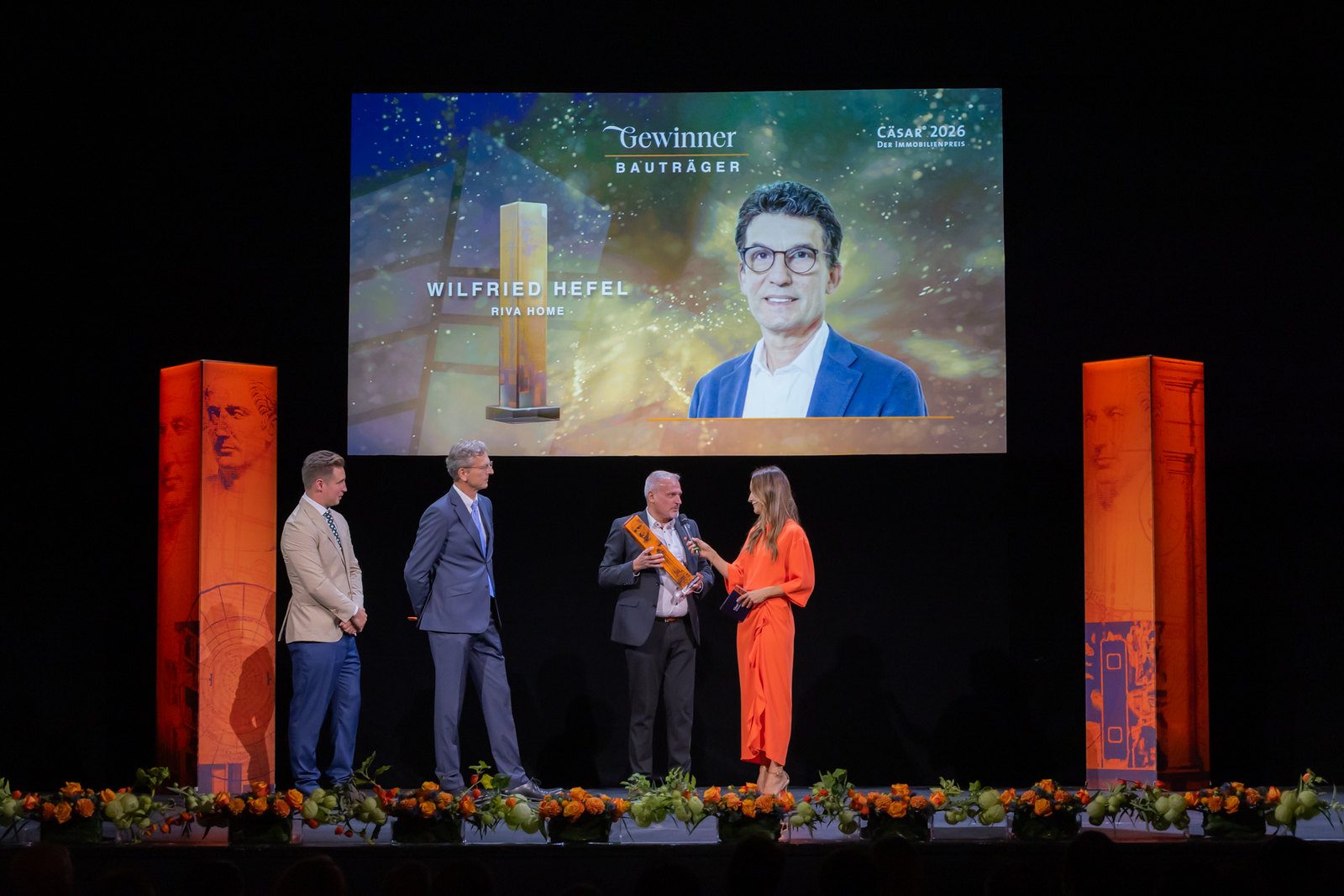 Bühne der CÄSAR® Gala in Wien mit orangen Award-Säulen; auf der Leinwand wird Wilfried Hefel, RIVA home, als Gewinner in der Kategorie Bauträger gezeigt