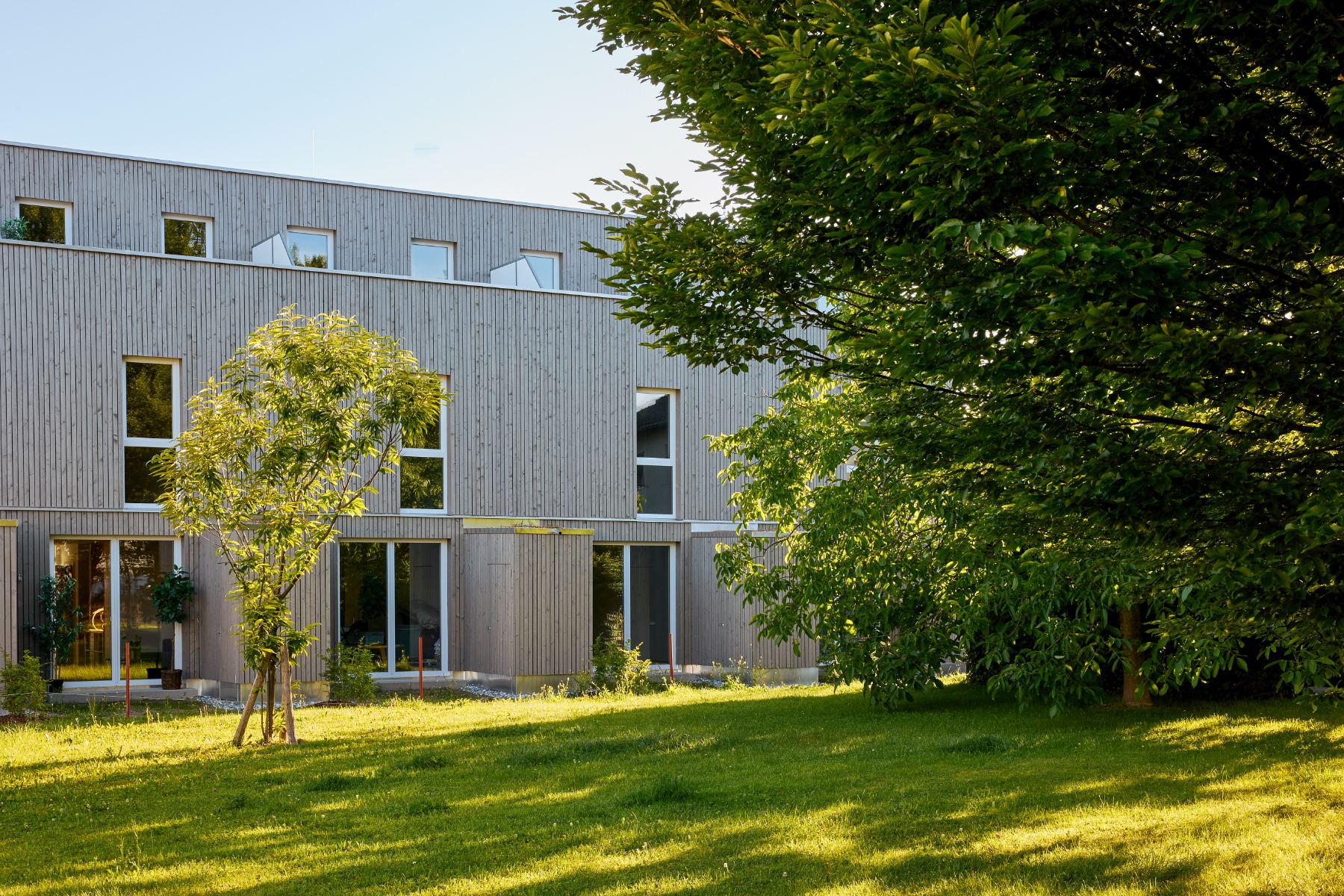 RIVA Wohnanlage mit grauer Holzfassade, großen Fenstern und Garten im Abendlicht